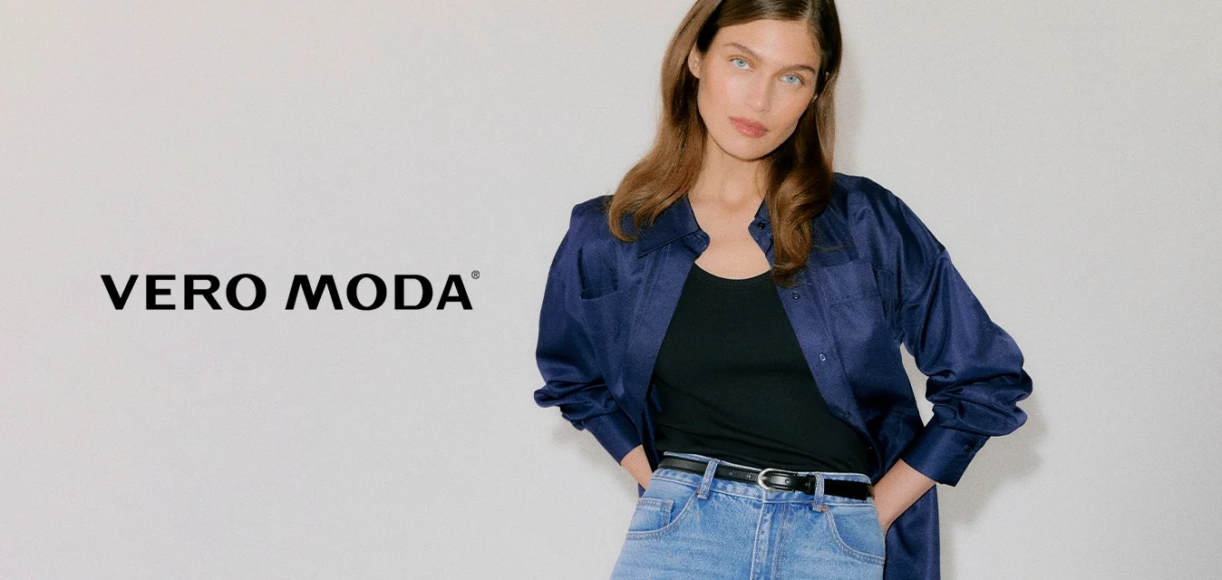 Giovane donna che indossa una camicia di raso blu scuro sopra una canottiera nera, jeans a vita alta azzurri e tacchi neri con occhielli.