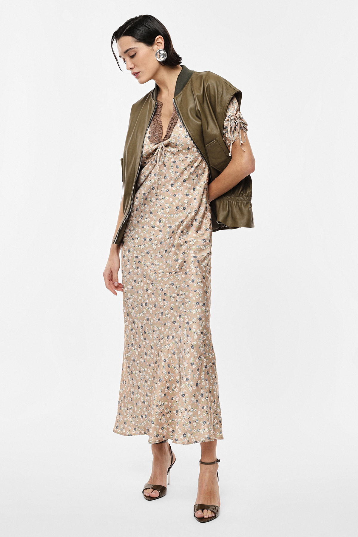 Woman in a beige floral slip dress with olive bomber jacket and heels, posing against a plain white background