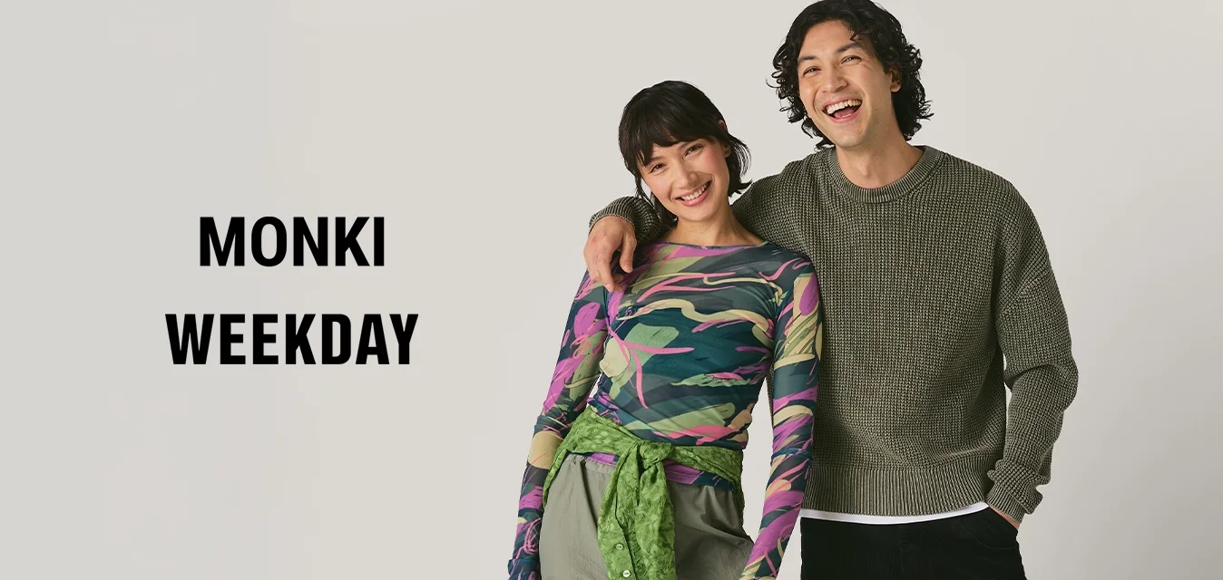 Un giovane uomo e una giovane donna stanno vicini, sorridendo; la donna indossa un top a motivi e pantaloni verdi, tenendo una borsa nera, mentre l'uomo indossa un maglione textured e pantaloni neri.