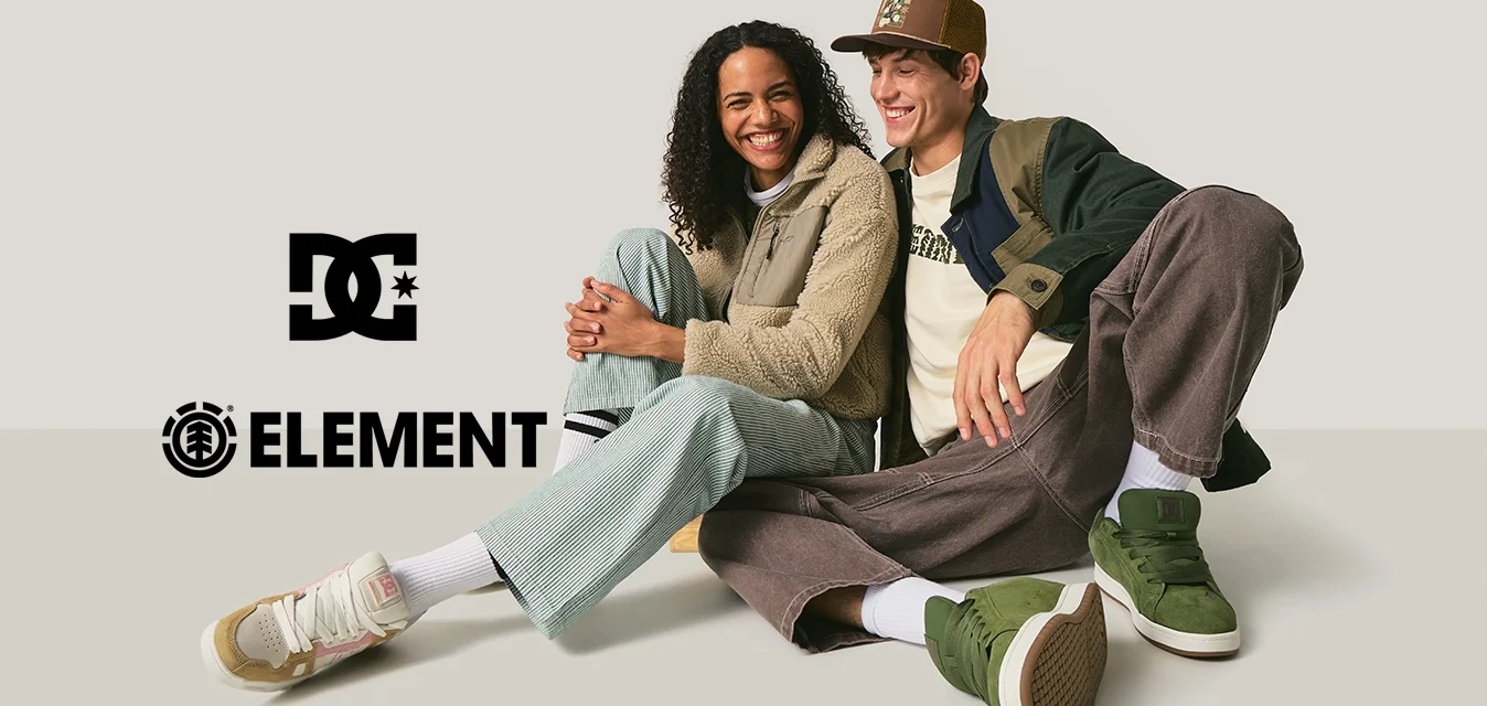 Due persone sedute a terra, indossano una giacca in pile beige, pantaloni verdi in velluto a coste e scarpe da ginnastica verdi, con un berretto marrone e una giacca blu.