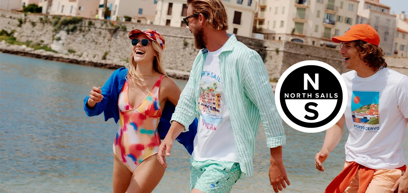 Tre amici in abiti estivi colorati camminano e ridono lungo un lungomare con edifici storici sullo sfondo sotto un cielo sereno.