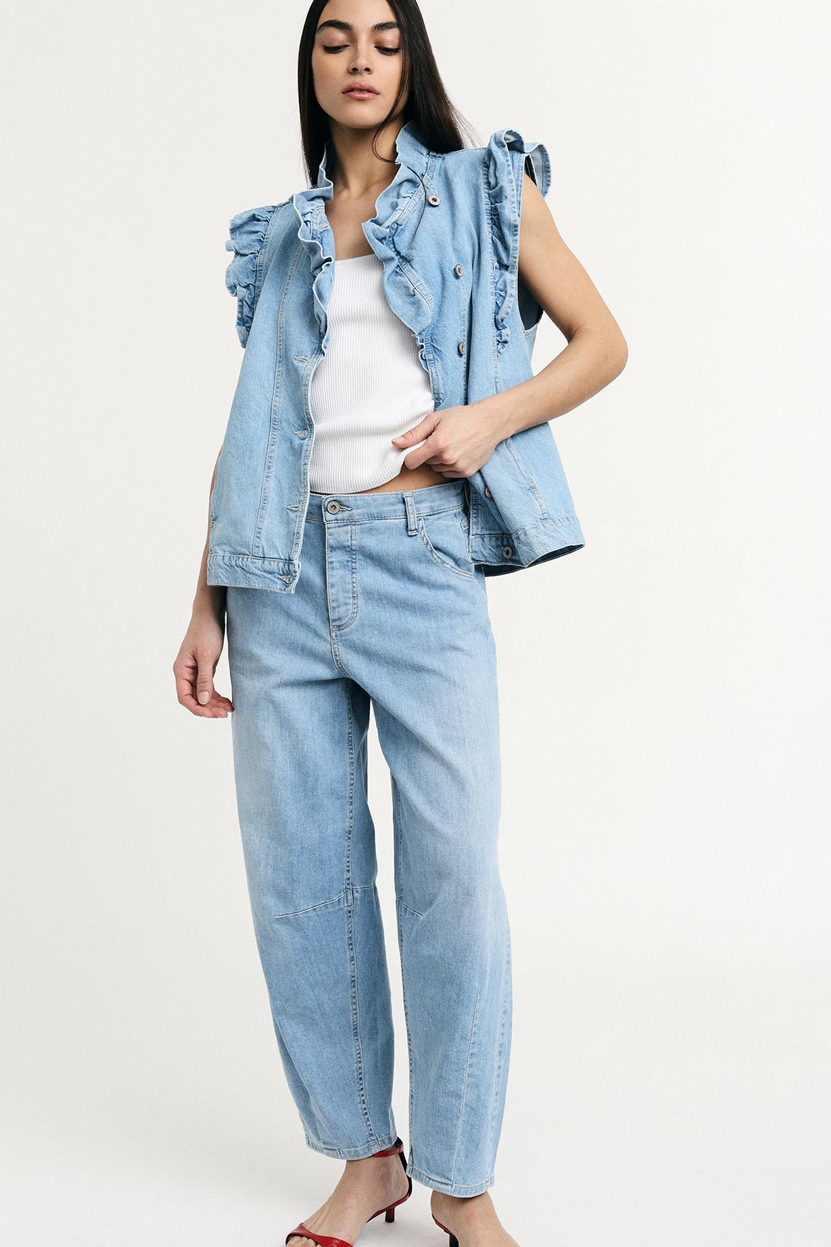 Woman wearing light-blue ruffled sleeveless denim jacket, white ribbed tank and high-waist straight light-blue jeans with red heels