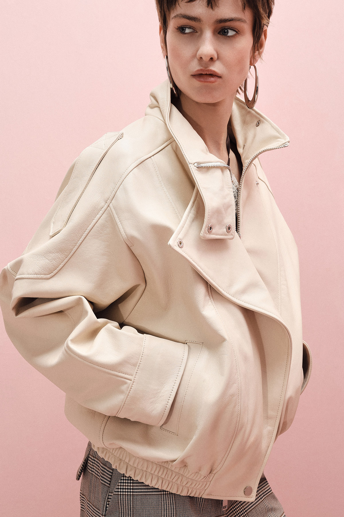 Young person in oversized cream leather jacket with hoop earrings and checked trousers against pink backdrop