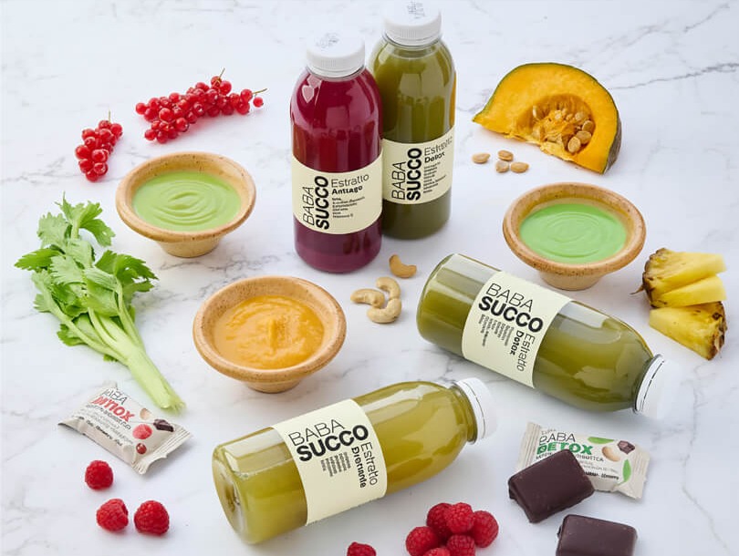 composizione di bottiglie babasucco con estratti colorati affiancate da verdure, frutta secca e creme vegetali su piano chiaro