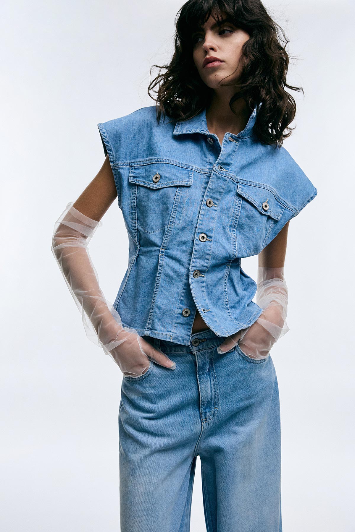 Woman wearing sleeveless denim button-up waist-cinched jacket and high-waisted jeans with translucent tulle gloves on a plain studio background