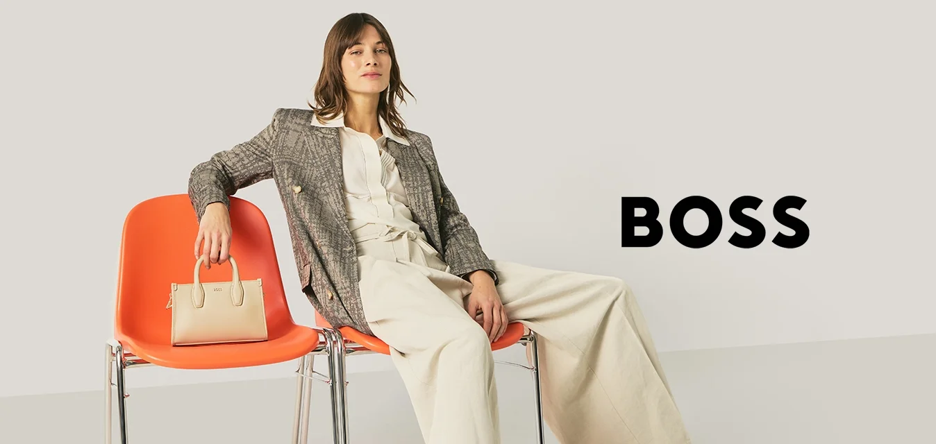 Donna in abito da lavoro seduta in modo rilassato su due sedie arancioni, con una borsa beige appoggiata su una delle sedie, su uno sfondo chiaro e uniforme.