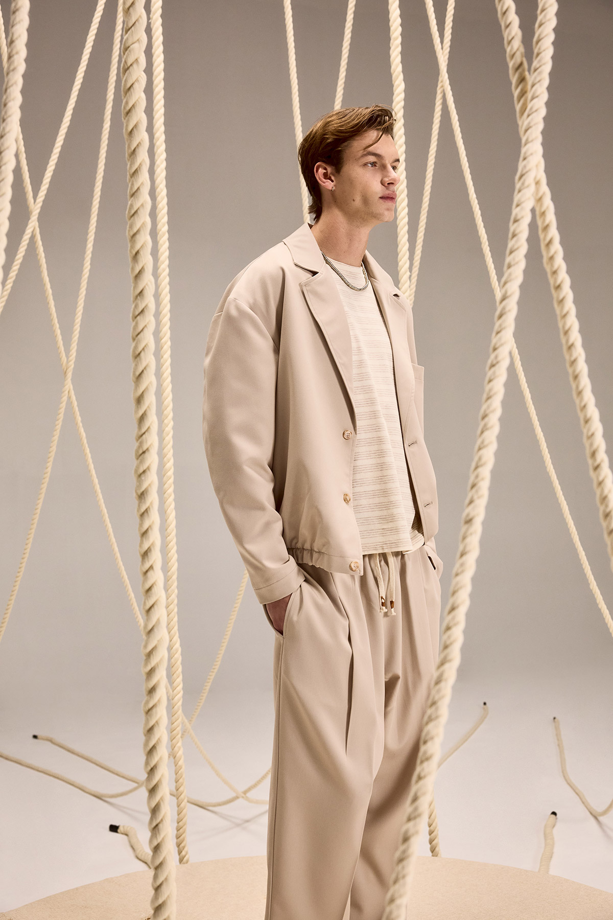 Male model in beige relaxed suit and knitted top standing among hanging thick ropes in a minimalist studio
