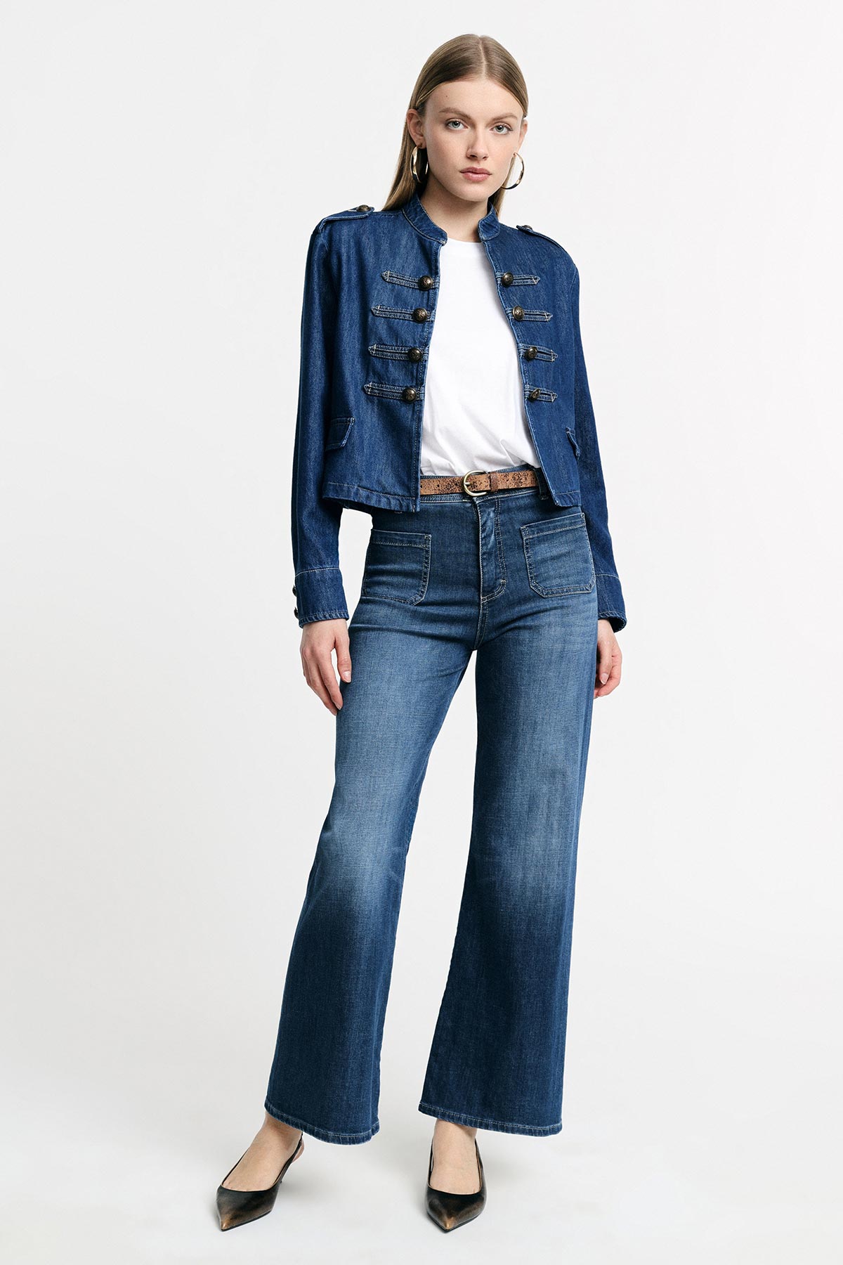 Woman modelling denim military-style jacket, white T-shirt, high-waisted wide-leg jeans, brown belt and black heels on plain background