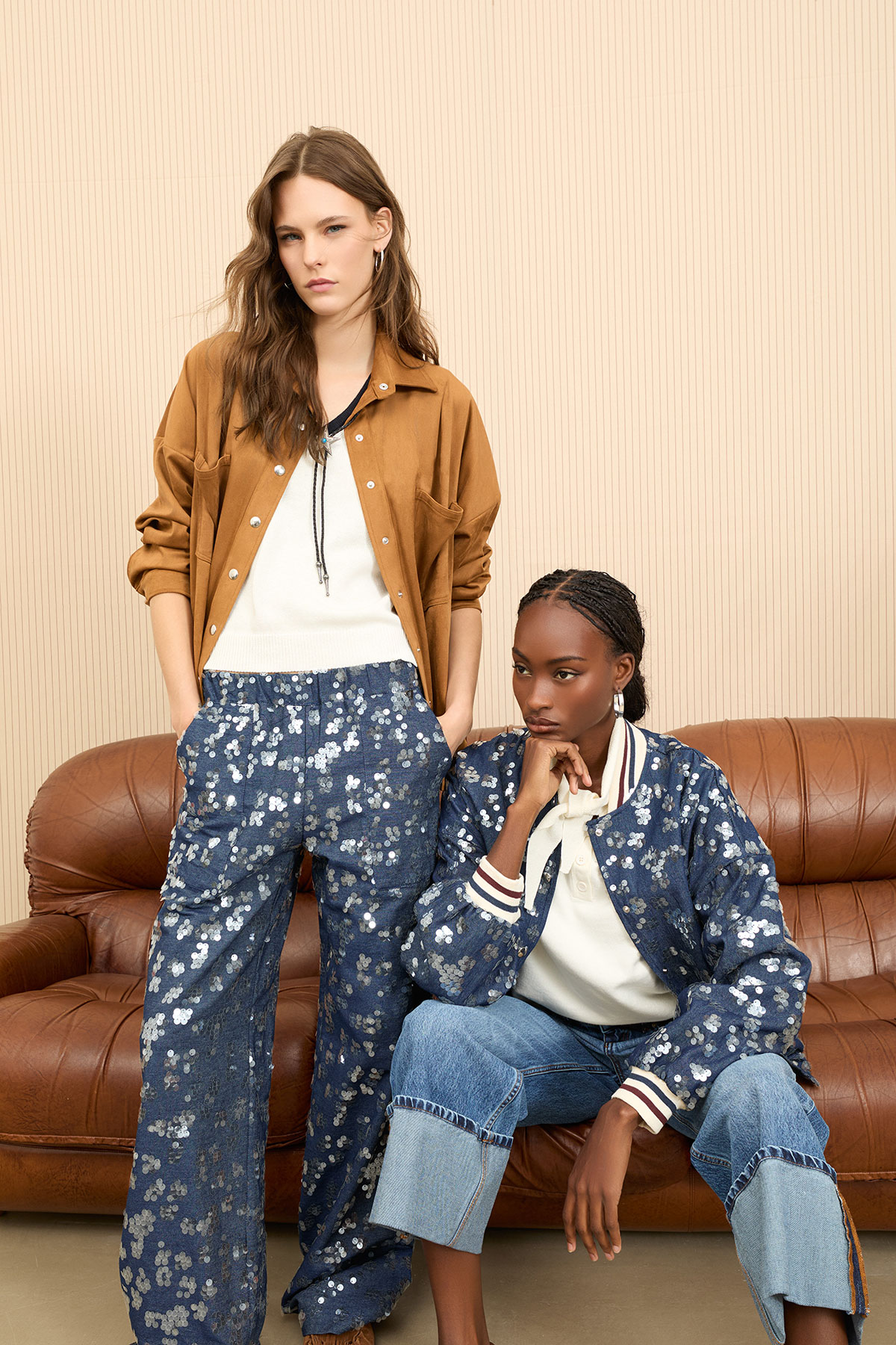 Two models in floral-patterned denim outfits — one standing in a brown shirt, one seated on a leather sofa against a neutral studio backdrop