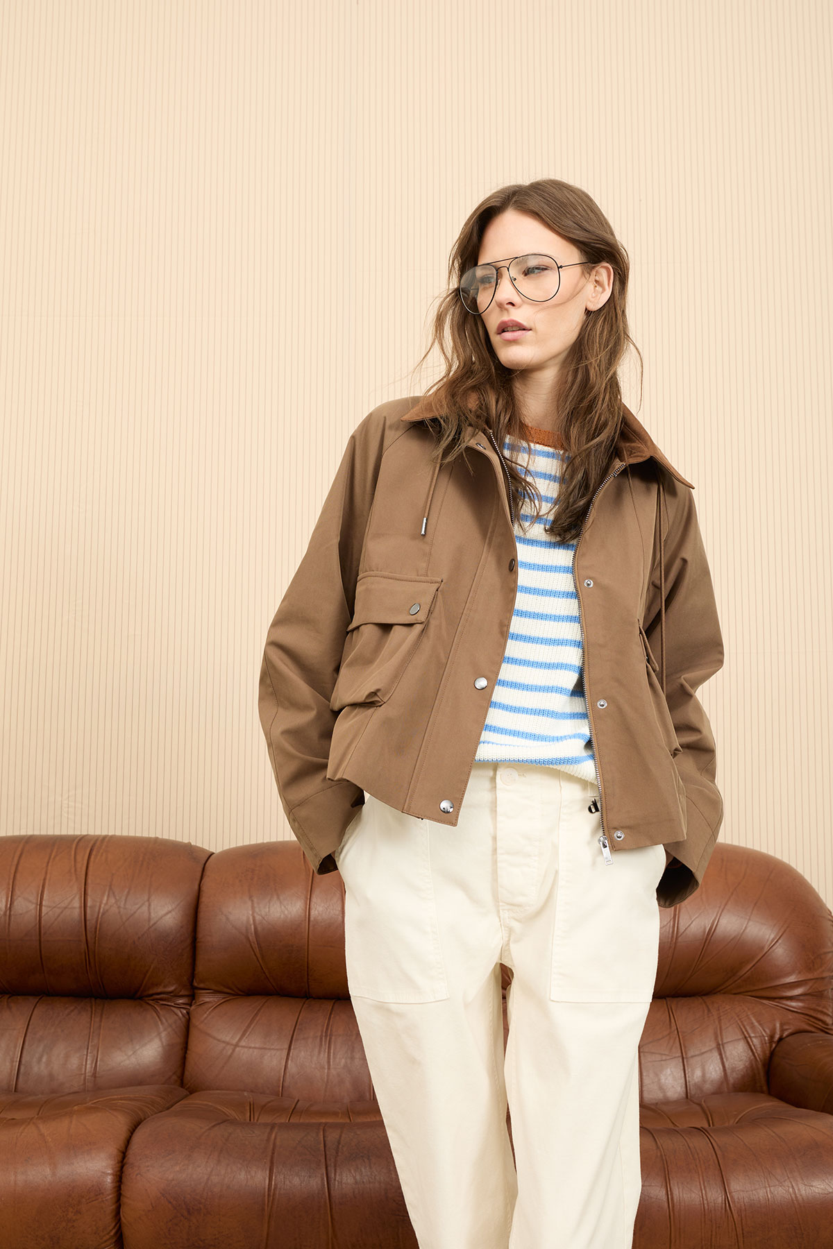 Woman wearing brown jacket, striped blue-and-white sweater and cream trousers standing in front of a brown leather sofa