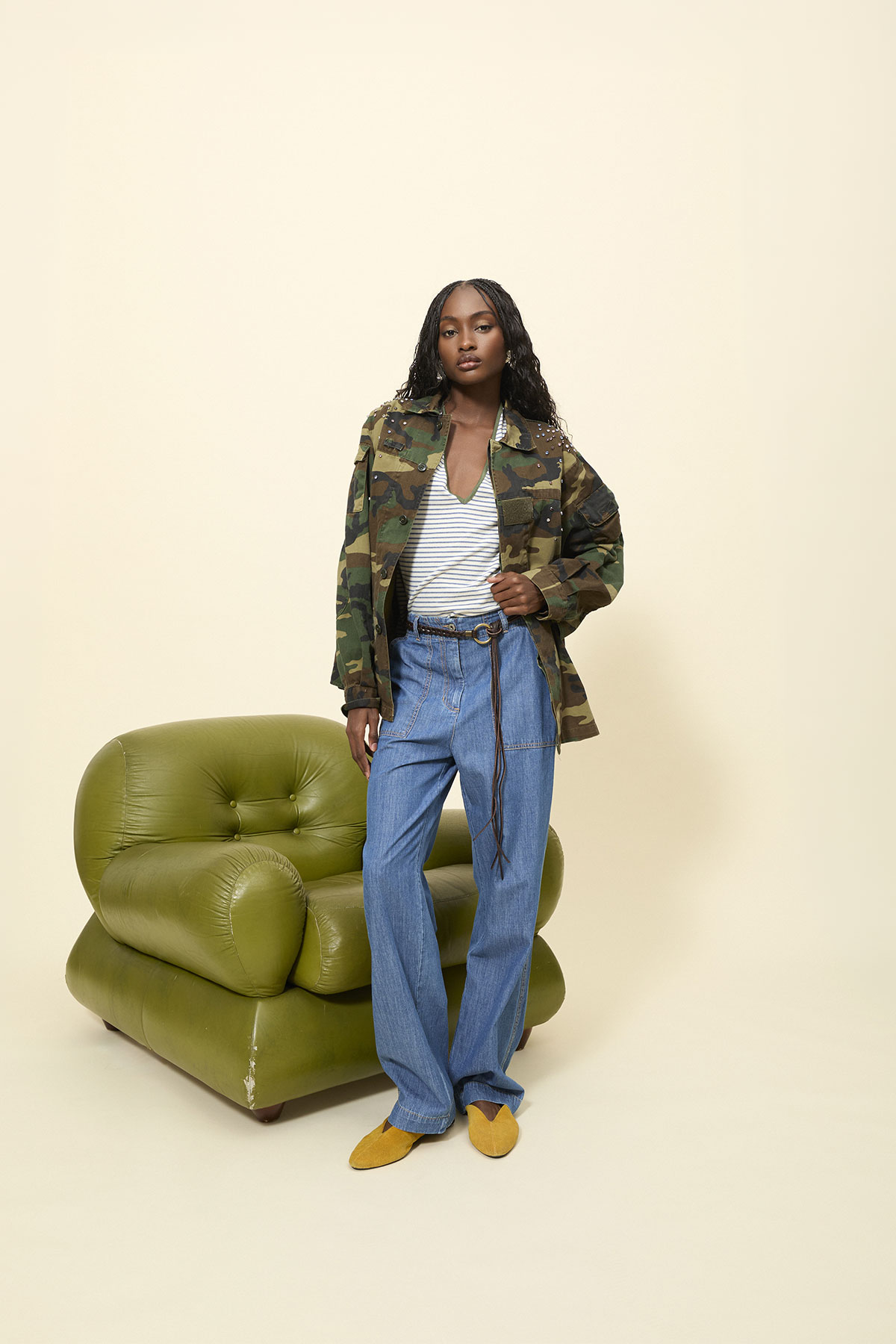 Person standing beside olive-green leather armchair wearing camouflage jacket, striped top, wide-leg jeans and mustard flats on cream background