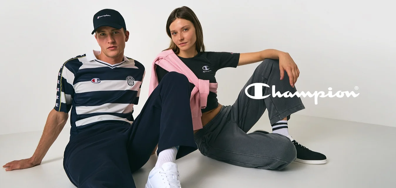 Un uomo e una donna sono seduti per terra contro una parete bianca, entrambi indossano abbigliamento casual Champion e mantengono pose rilassate.