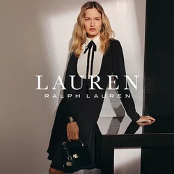 Donna con capelli biondi lunghi indossa un vestito nero con colletto bianco a bavero, tiene una borsa nera in mano, in piedi accanto a un tavolo scuro lucido.