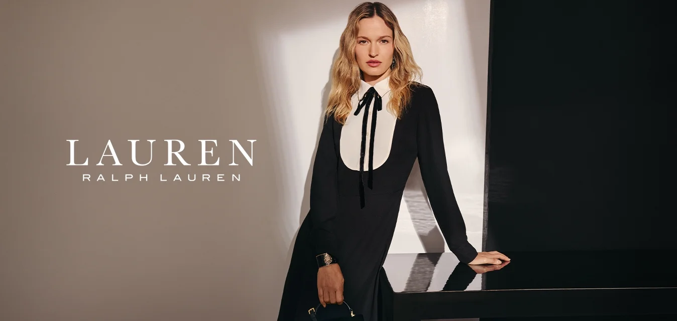 Donna con capelli biondi lunghi indossa un vestito nero con colletto bianco a bavero, tiene una borsa nera in mano, in piedi accanto a un tavolo scuro lucido.