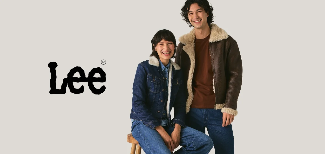 Due persone che indossano giacche di jeans e shearling; una è seduta su uno sgabello mentre l'altra sta in piedi, entrambe sorridenti contro uno sfondo semplice.