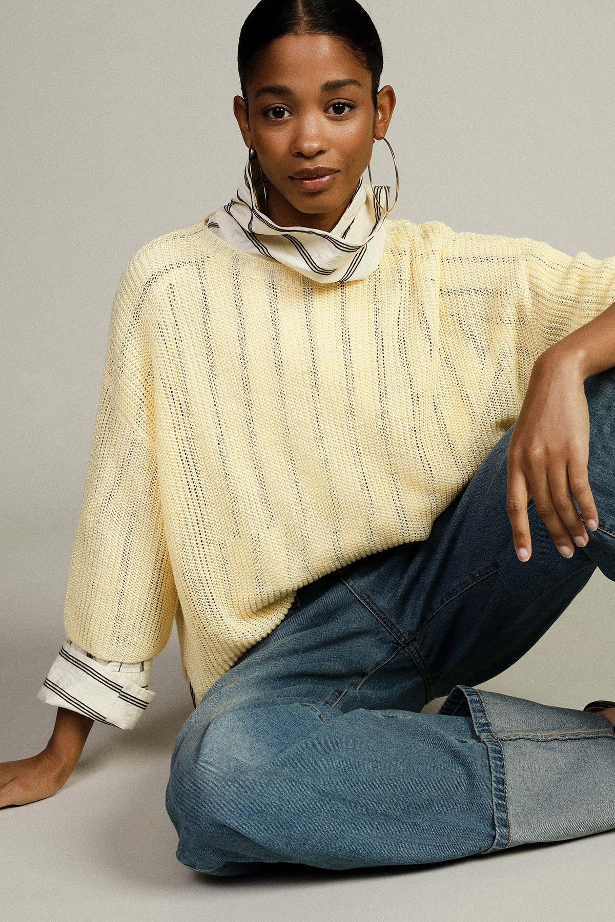 Model wearing pale yellow knitted sweater, striped neck scarf, hoop earrings and relaxed blue jeans pose against neutral background