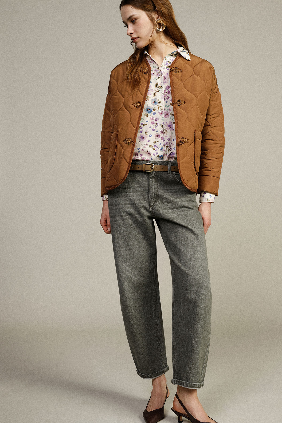 Woman modelling brown quilted jacket over floral blouse with grey high-waist jeans, slingback heels and gold hoop earrings on neutral background