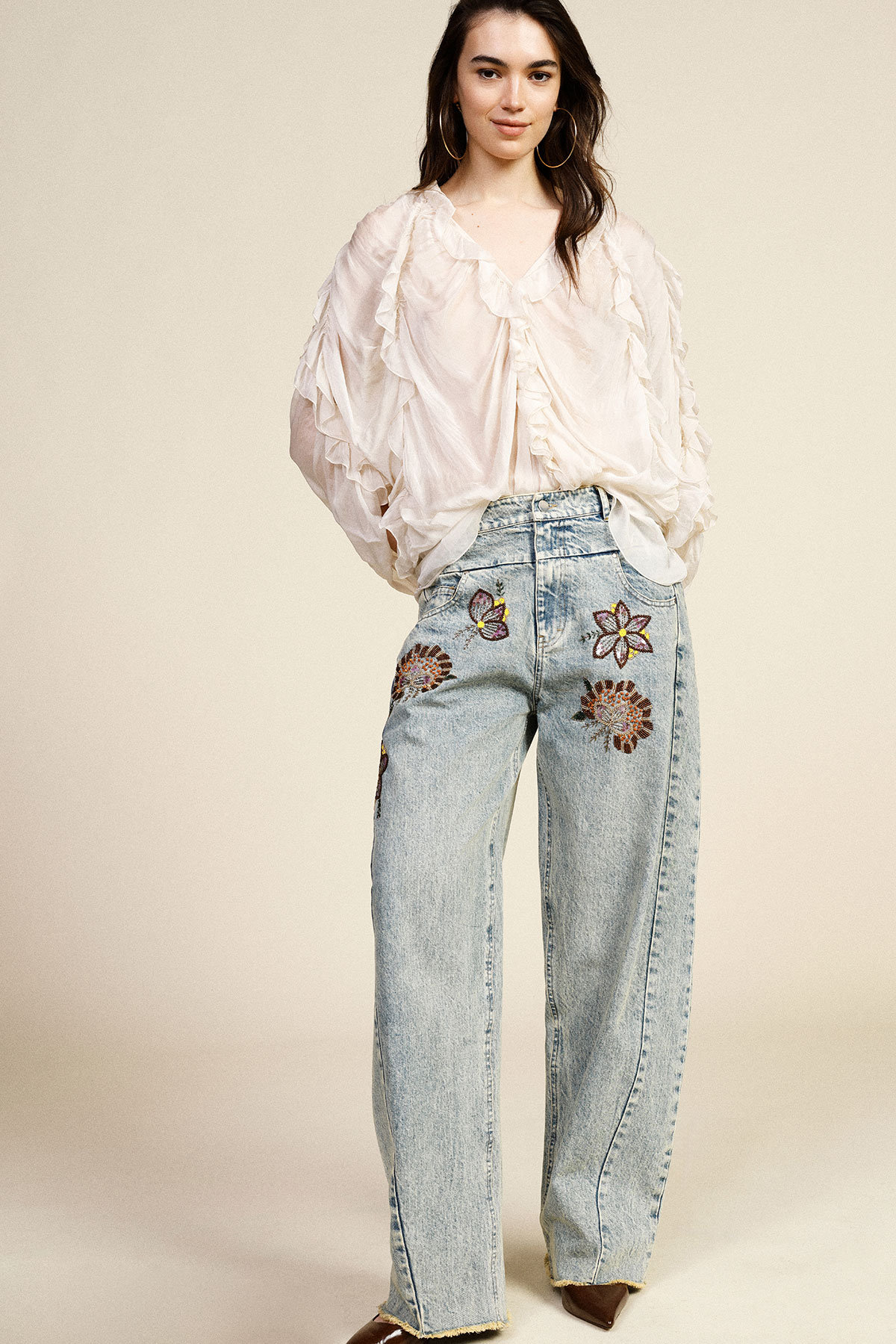 Woman wearing sheer ruffled blouse and light denim jeans embroidered with flowers and butterfly against a neutral background