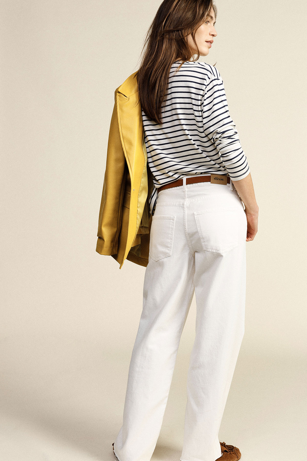 Woman wearing navy-striped long-sleeve top, white wide-leg jeans and brown loafers, yellow blazer draped over shoulder, posing back