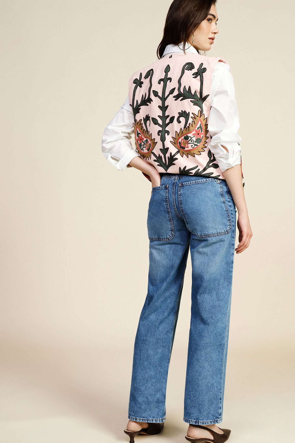 Woman standing with back to camera wearing an embroidered pink waistcoat over a white shirt and high-waisted straight blue jeans