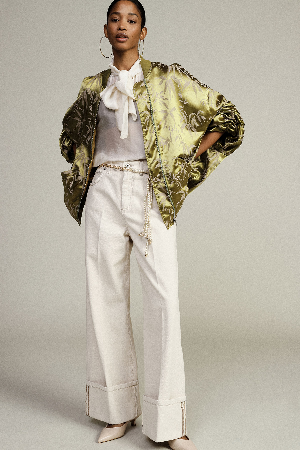 Model wearing green satin floral bomber jacket, white bow blouse, wide-leg cream trousers and heels