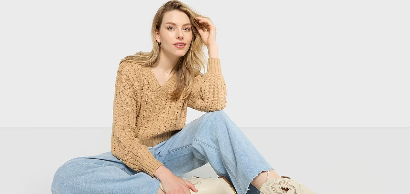 Donna con lunghi capelli biondi seduta con le gambe incrociate, indossa un maglione di lana beige, jeans strappati azzurri e stivali da cowboy ricamati di color crema.