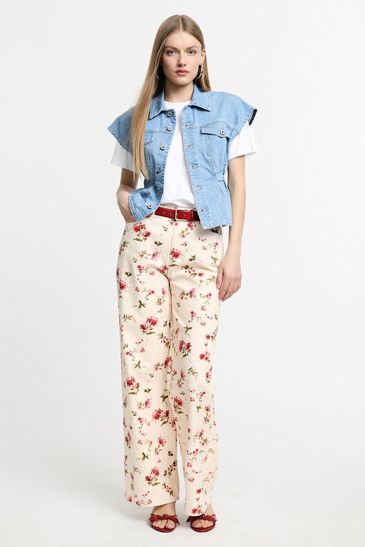 Woman wearing cropped denim vest over white T-shirt, floral wide‑leg trousers with red belt and sandals, standing on white background