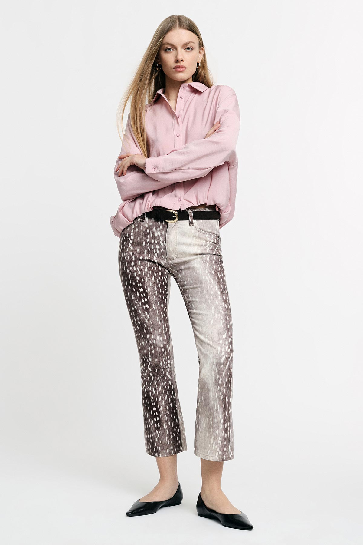 Young woman model wearing a pink blouse, patterned cropped trousers with a black belt and black flats, standing