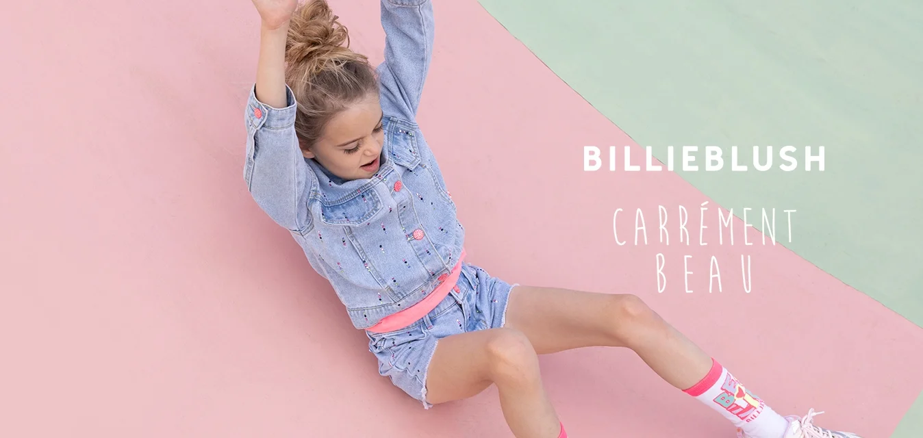 Ragazza giovane con giacca di jeans e pantaloncini che scivola lungo una superficie da gioco curva rosa e verde con le braccia sollevate.