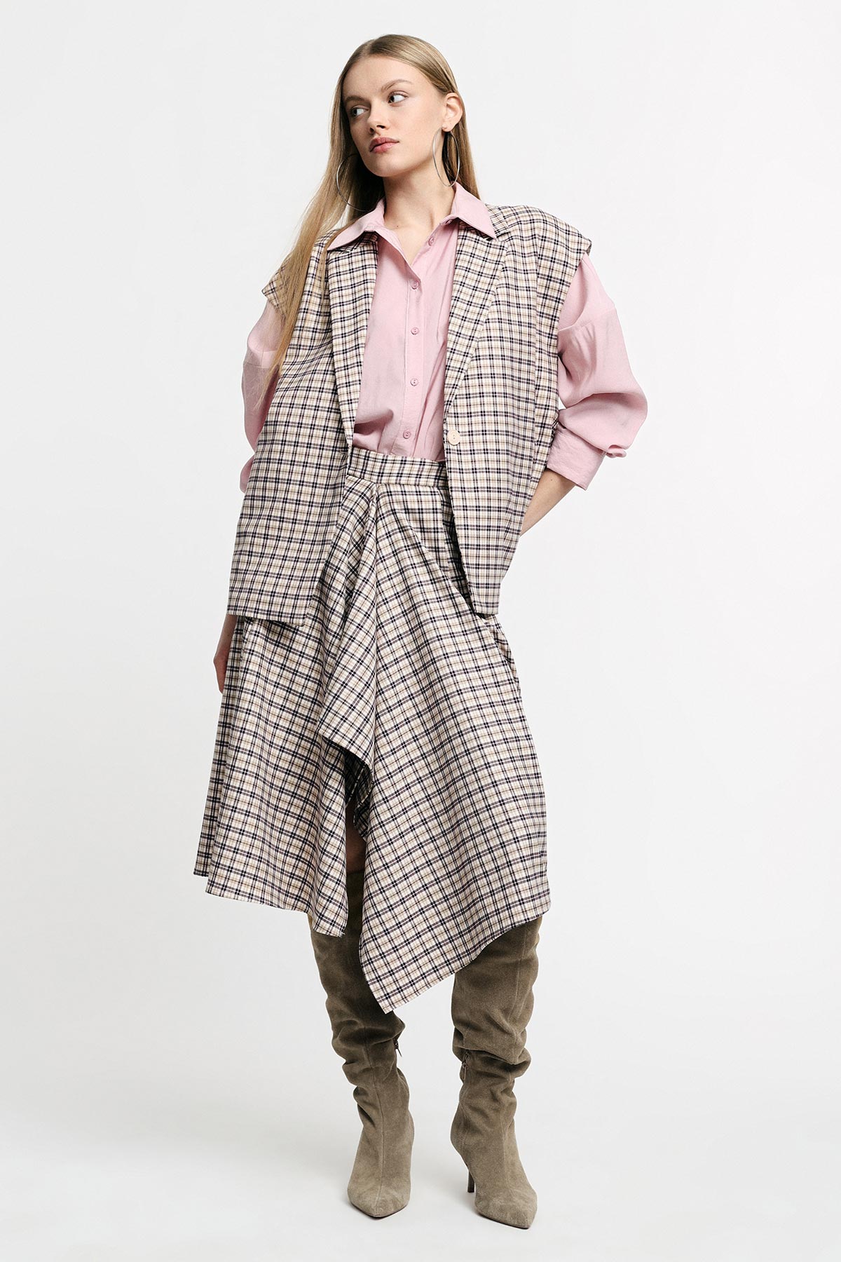 Woman standing in a pink blouse, checked oversized vest and asymmetric plaid skirt paired with taupe suede knee-high boots