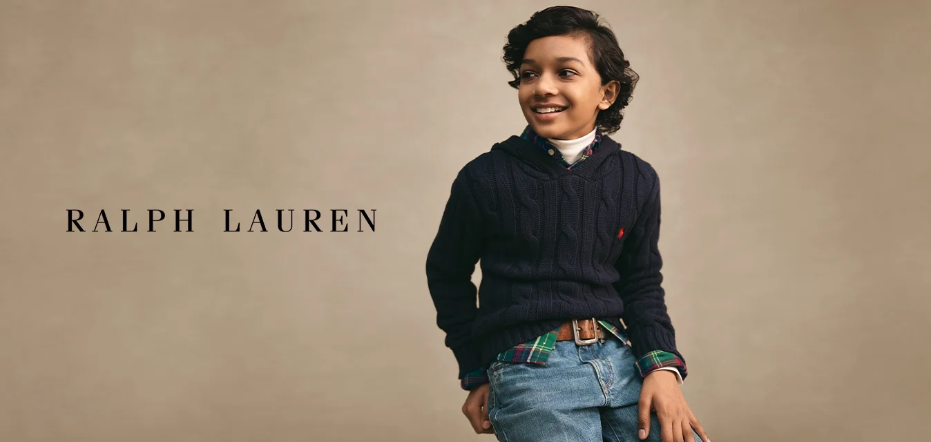 Ragazzo con capelli scuri e ricci che indossa un maglione blu navy sopra una camicia a quadri e jeans, sorridente e con lo sguardo rivolto di lato su uno sfondo neutro.