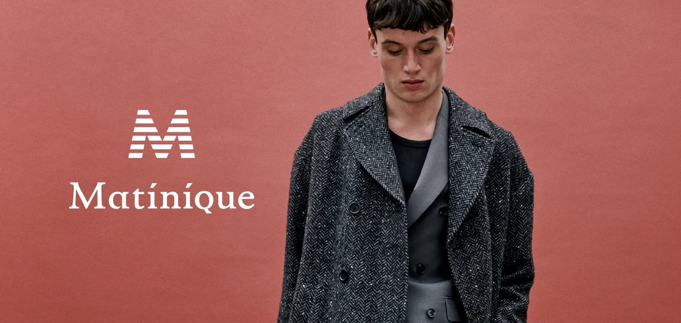 Uomo in abito grigio e soprabito a motivo, in piedi su pavimento di cemento davanti a uno sfondo color ruggine, con le mani in tasca.