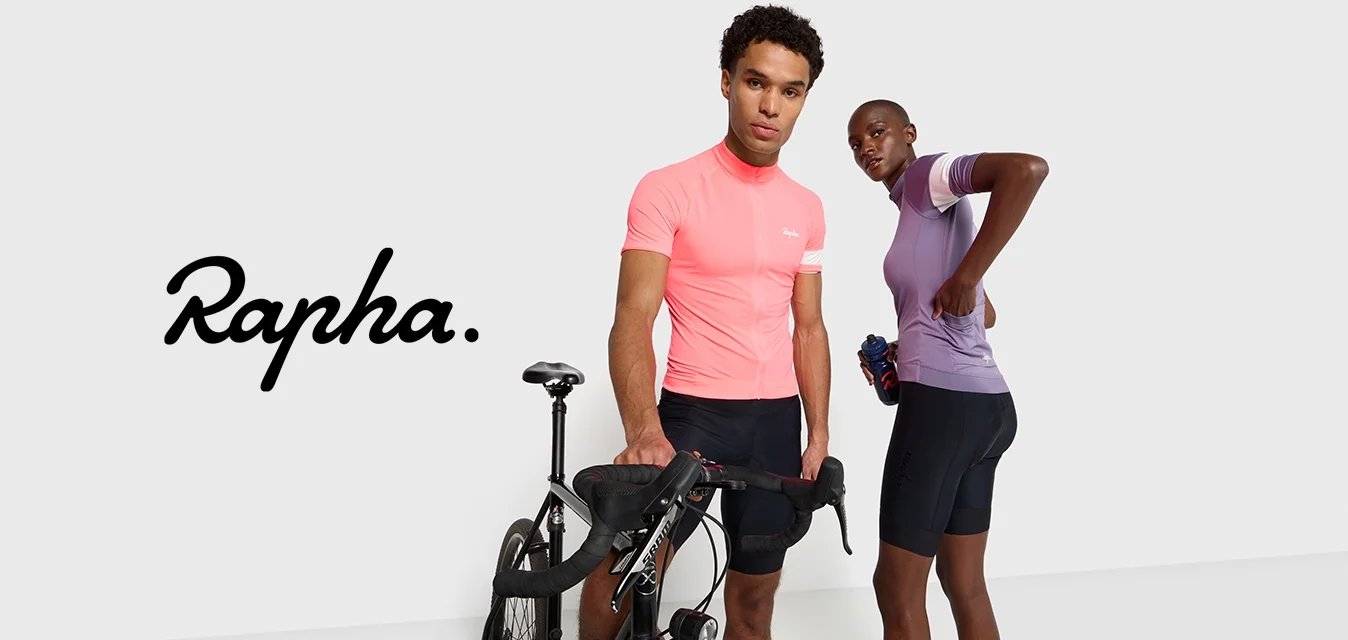 Due persone stanno in piedi accanto a una bicicletta. Una indossa una maglietta da ciclismo aderente rosa e pantaloni neri, mentre l'altra indossa una maglietta viola e pantaloni scuri.