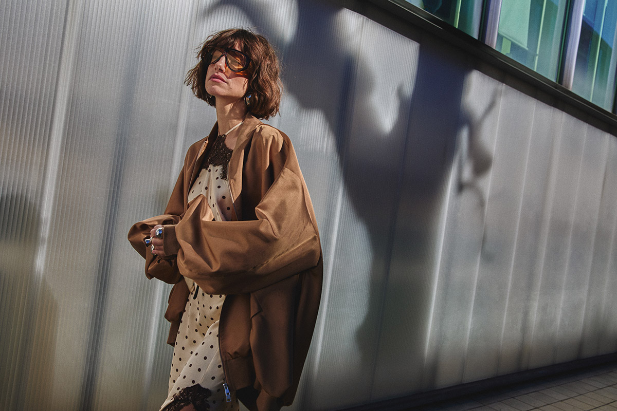 Fashionable woman in oversized brown jacket and polka-dot dress wearing sunglasses against corrugated metal wall with dramatic shadow