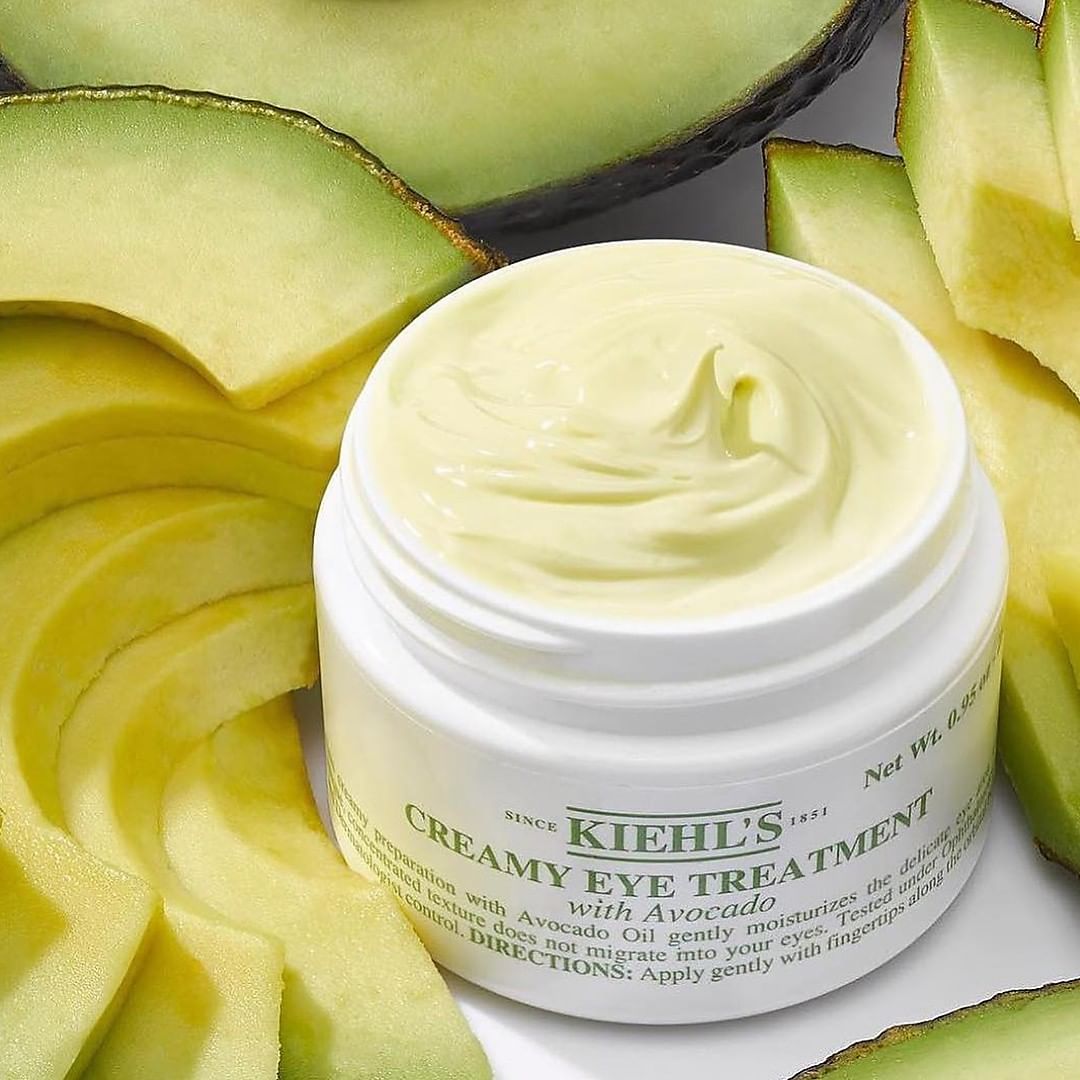 Vasetto aperto di Kiehl&rsquo;s Creamy Eye Treatment with Avocado con texture visibile, circondato da fette di avocado fresco su sfondo verde.