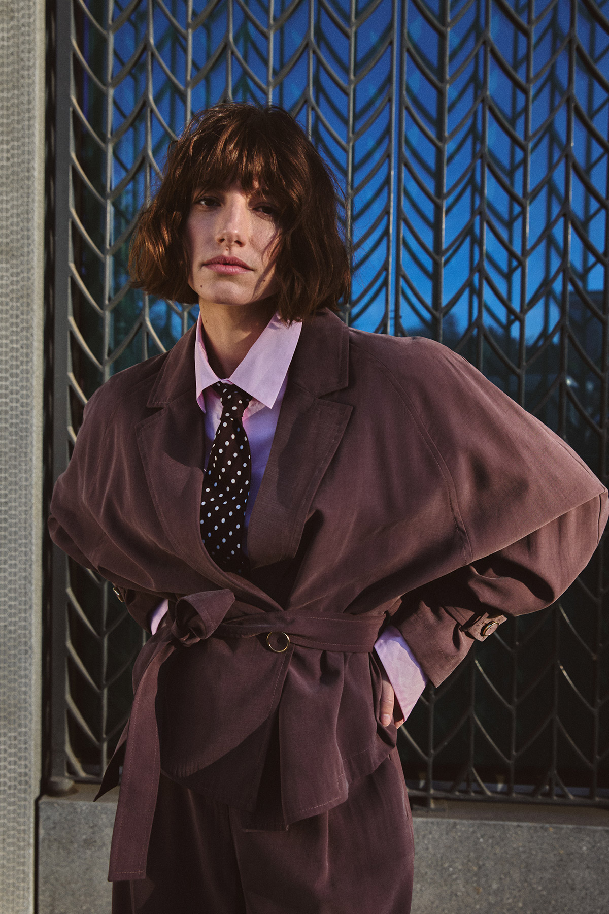 Person wearing an oversized mauve suit, pink shirt and polka-dot tie standing against a patterned metal gate