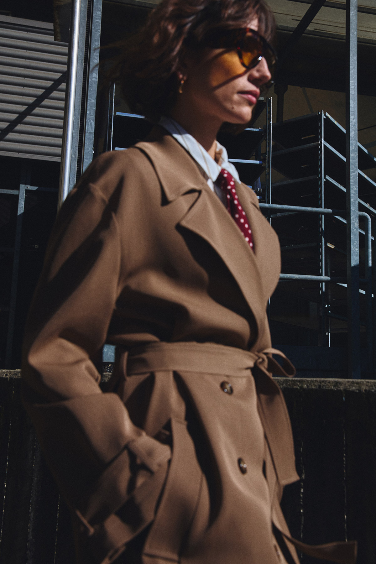 Person wearing a tan trench coat, red polka-dot tie and sunglasses, walking past metal industrial scaffolding in urban setting