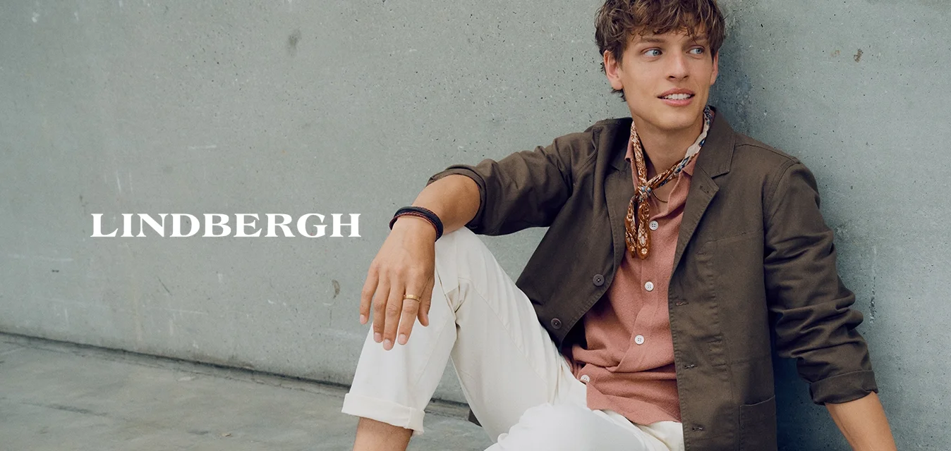 Giovane uomo seduto sul pavimento contro un muro di cemento, indossa una giacca marrone, camicia rosa, pantaloni bianchi, mocassini color cuoio e una sciarpa al collo.