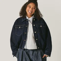 Persona sorridente in piedi, che indossa una giacca scura oversize, pantaloni larghi scuri e una camicia bianca, su uno sfondo chiaro e uniforme.