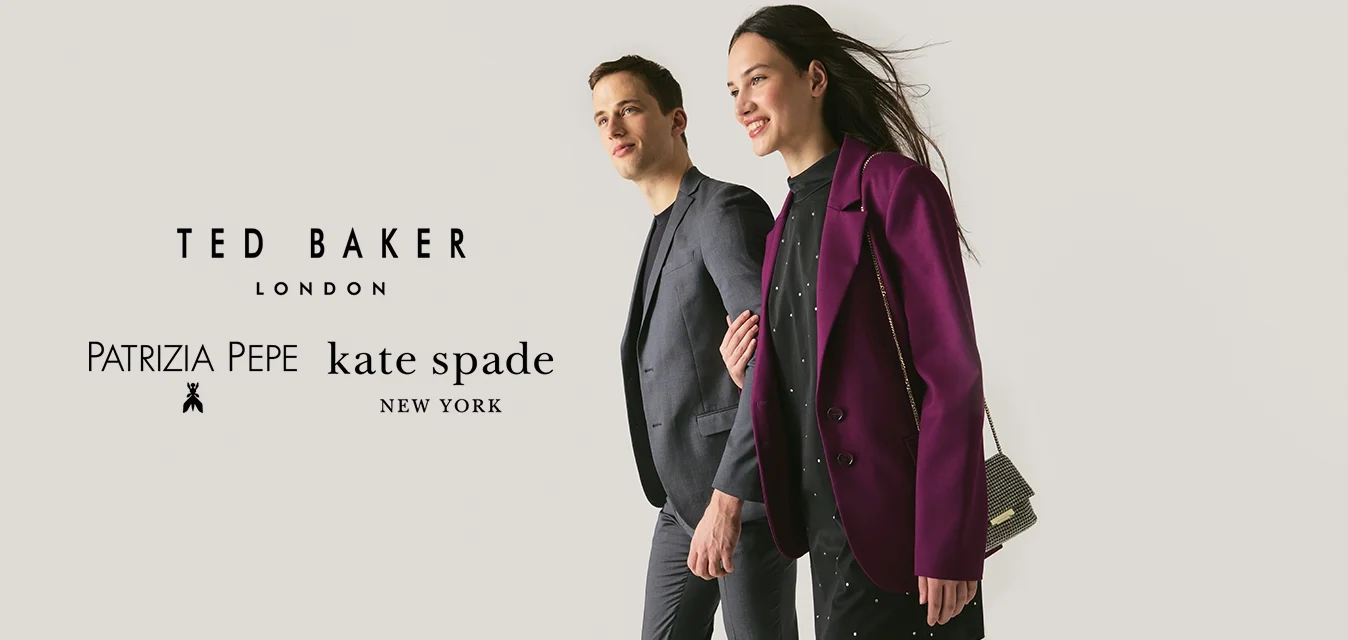 Uomo e donna che camminano fianco a fianco, donna con blazer viola e vestito nero che tiene una piccola borsa a catena, uomo in abito grigio scuro.