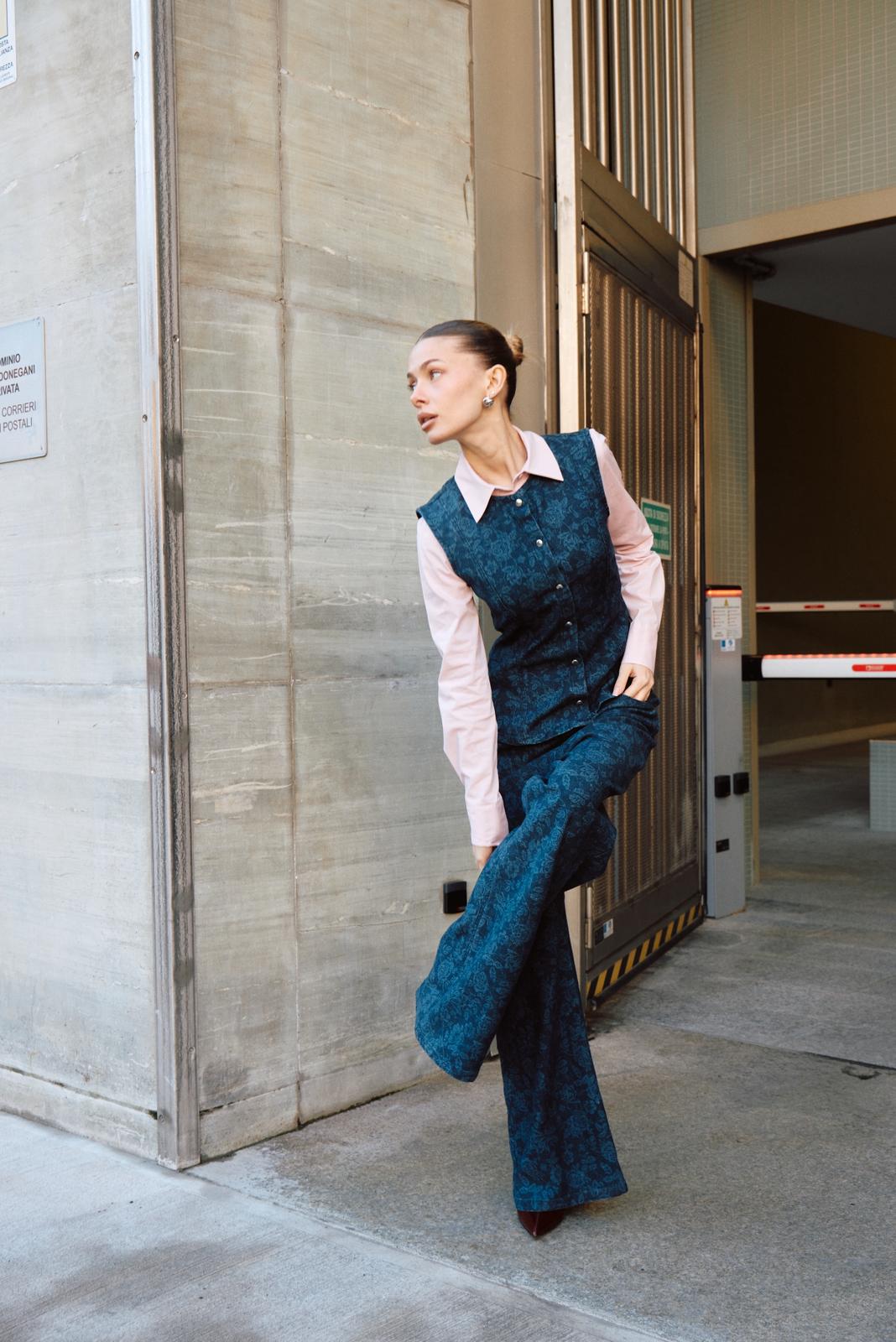 Woman in patterned navy tailored vest and flared trousers with pink shirt posing by industrial garage entrance in urban setting