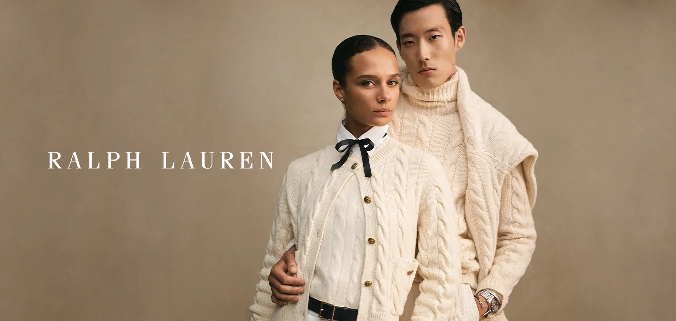 Due modelli che indossano maglioni a treccia color crema posano vicino su uno sfondo beige, uno con un papillon e l'altro con un orologio.