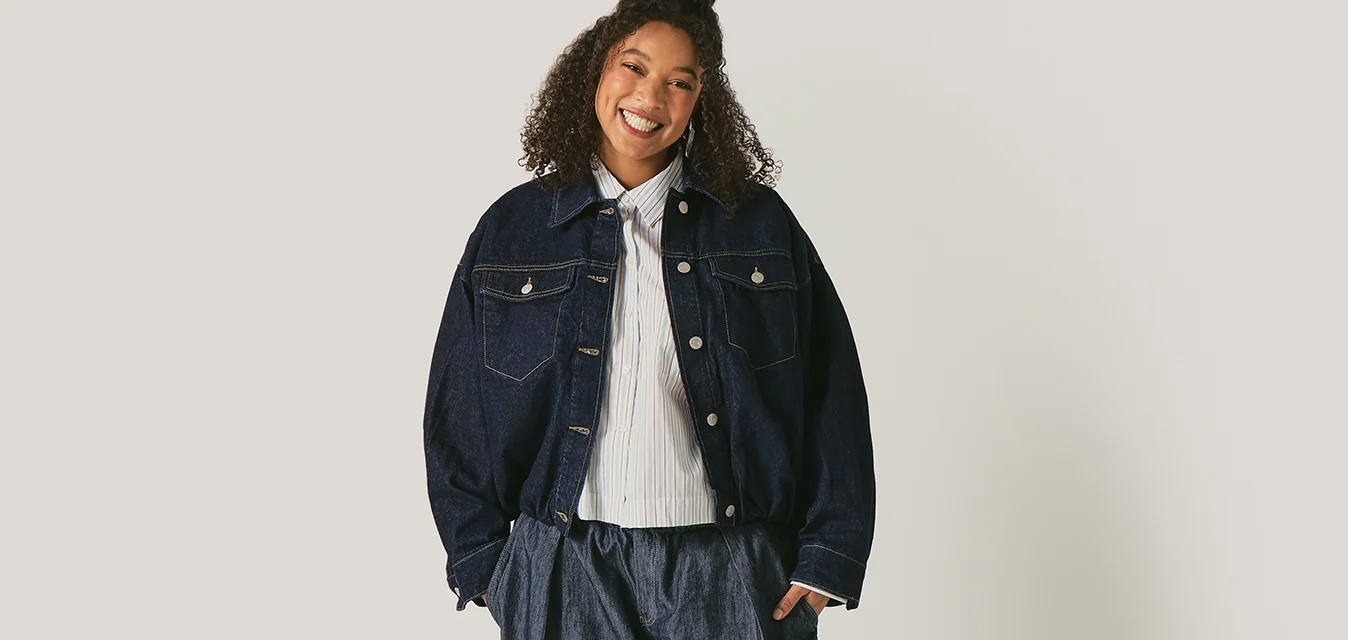 Persona sorridente in piedi, che indossa una giacca scura oversize, pantaloni larghi scuri e una camicia bianca, su uno sfondo chiaro e uniforme.