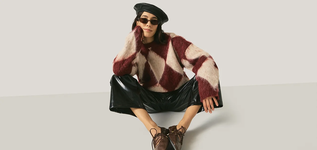 Donna con basco nero, occhiali da sole, maglione a motivi bordeaux e bianchi, pantaloni neri lucidi e scarpe marroni con punta, seduta con le gambe divaricate.