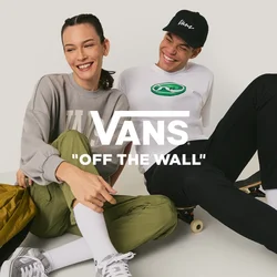 Due giovani seduti per terra in abiti casual, uno con uno skateboard e uno zaino giallo, sorridenti e vicini tra loro.