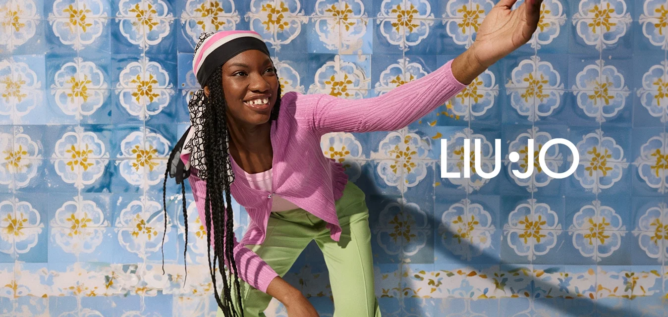 Cardigan a coste rosa, top rosa chiaro e pantaloni verdi con una texture liscia. Sullo sfondo, piastrelle blu con motivi floreali in giallo e bianco.