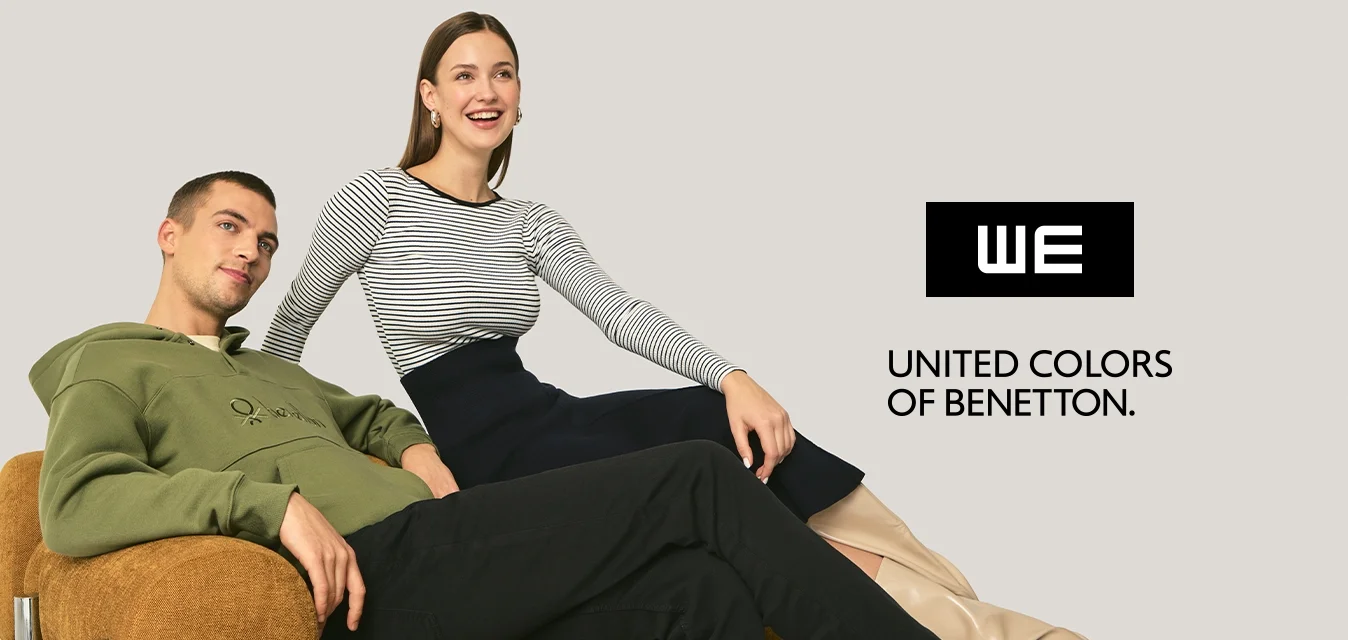 Felpa verde con cappuccio, top a righe, gonna nera e stivali beige. La modella è seduta su un divano marrone testurizzato con dettagli cromati, con uno sfondo neutro.