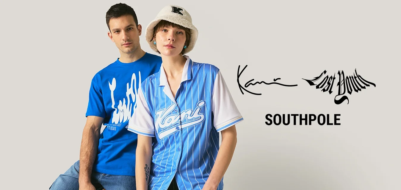 Giovane uomo con maglietta blu grafica e giovane donna con camicia a righe blu e cappello a secchiello bianco posano davanti a uno sfondo neutro.