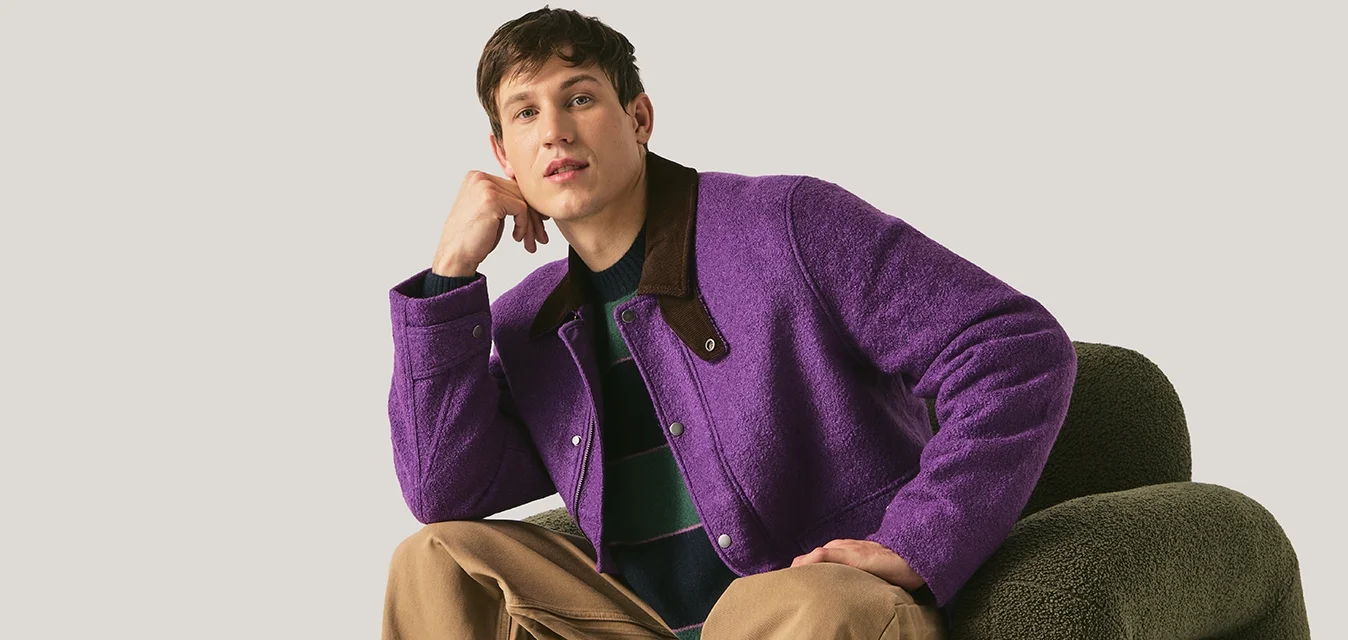 Giovane uomo con giacca viola e pantaloni beige seduto su una poltrona verde scuro dalla forma unica, su sfondo semplice.