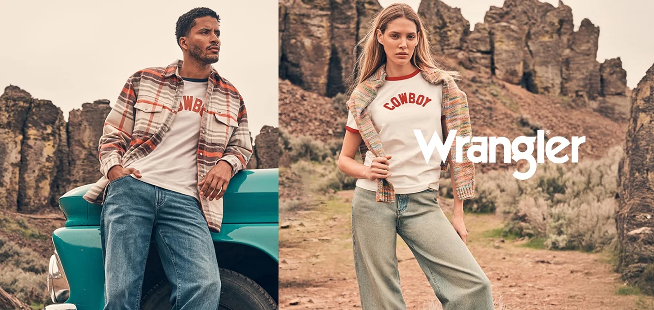 Un uomo si appoggia a un camion vintage turchese mentre una donna sta in piedi nelle vicinanze; entrambi indossano abiti casual a tema cowboy in un paesaggio desertico roccioso.